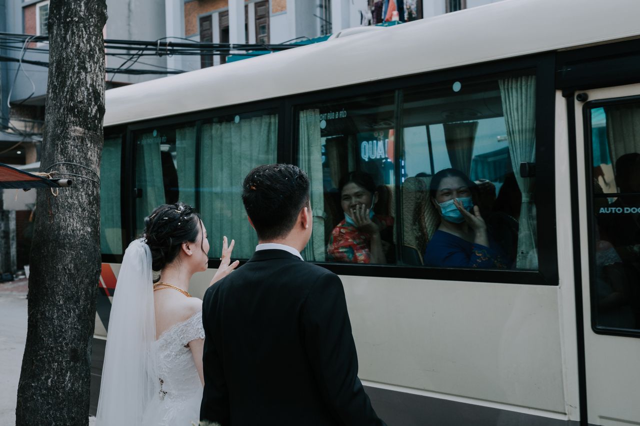 Young bride and groom saying goodbye and waving hands to guests in masks sitting in bus